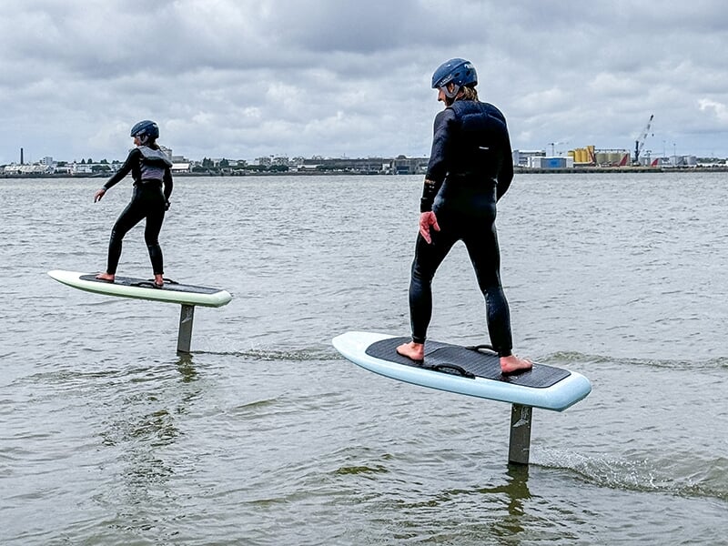 Deux personnes en combinaison et casque sur des planches de e-foil, lévitant au-dessus de l'eau, sous un ciel nuageux avec une ville en arrière-plan.