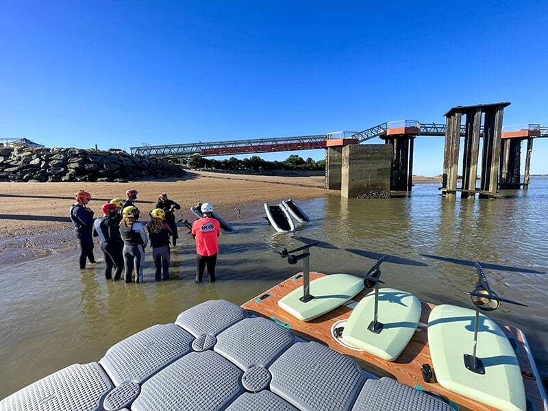Groupe de personnes debout dans l'eau près de la plage, discutant, avec des planches de e-foil et une structure en arrière-plan.