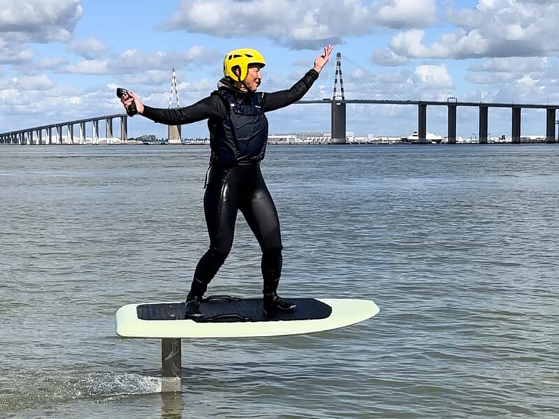 Personne en combinaison et casque jaune, les bras levés, debout sur une planche de e-foil, avec un grand pont visible à l'arrière-plan.