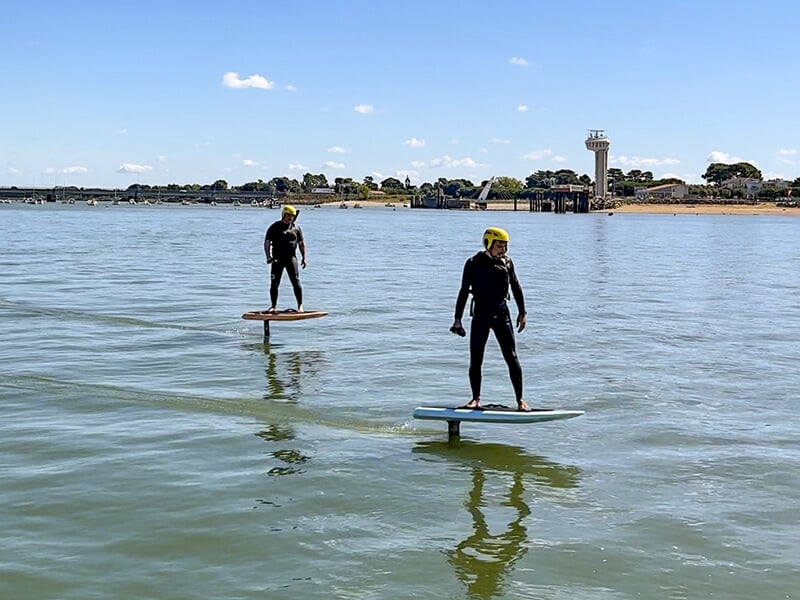 Deux personnes en combinaison et casque jaune faisant du e-foil sur une mer calme, avec des bâtiments et des structures en arrière-plan.