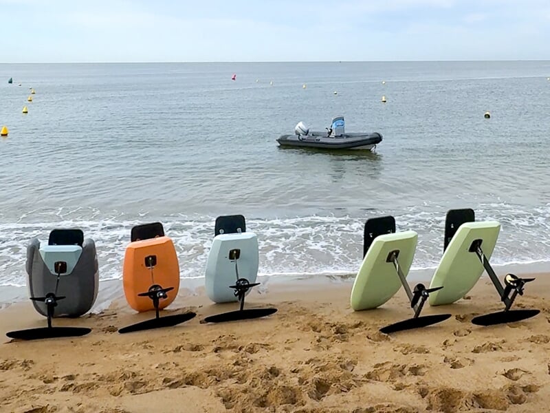 Cinq planches de e-foil alignées sur une plage, prêtes pour une session, avec un bateau pneumatique flottant à proximité.