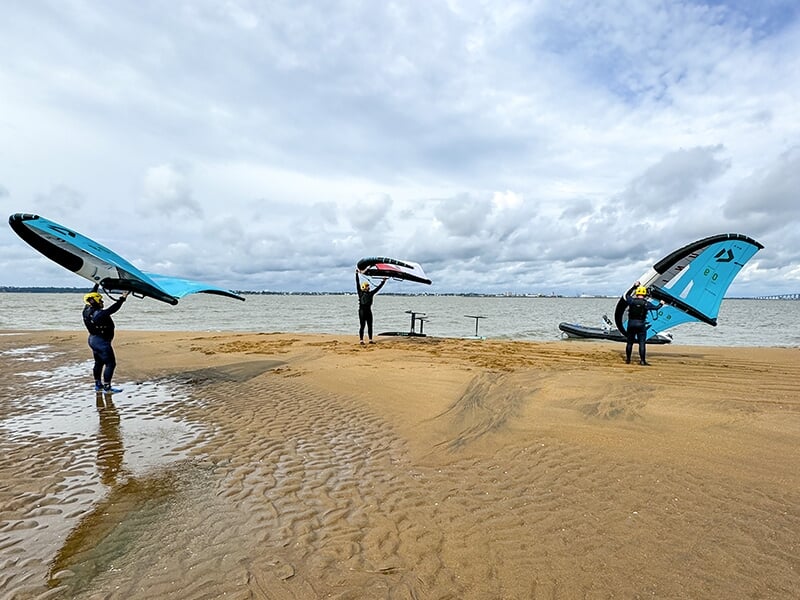 Trois surfeurs debout sur une plage de sable, tenant des ailes de wing foil, se préparant à naviguer.