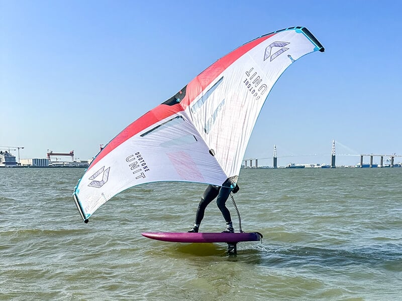 Surfeur utilisant une planche à foil et tenant une aile gonflable, glissant sur l'eau, sous un ciel dégagé.