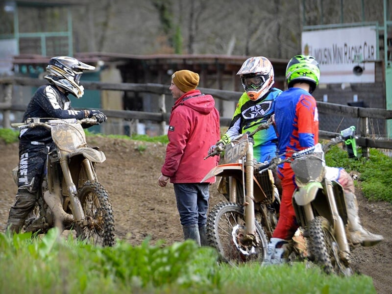 Trois motocyclistes en tenue de motocross, recouverts de boue, discutant avec un homme en veste rouge et bonnet, sur un circuit de motocross boueux