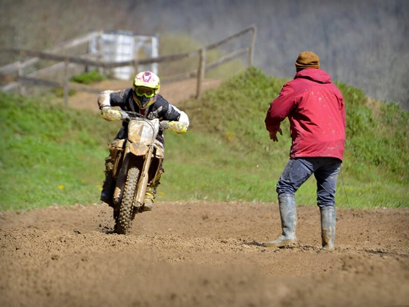 Un motocycliste roulant sur un terrain de motocross boueux, avec un homme en veste rouge et bonnet lui donnant des instructions depuis le bord de la piste.