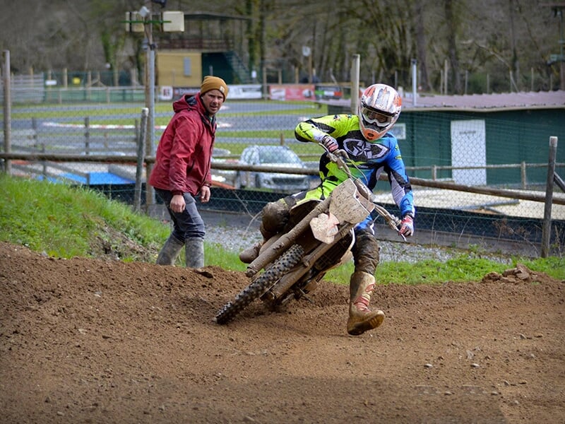 Motocycliste en pleine action dans un virage serré sur un terrain de motocross boueux, tandis qu'un homme en veste rouge se tient à côté en observant.