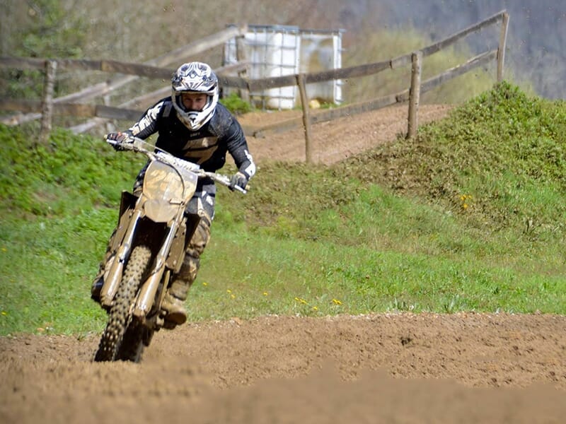 Motocycliste en combinaison noire et blanche roulant rapidement sur une piste de motocross boueuse, entouré de verdure et de barrières en bois.