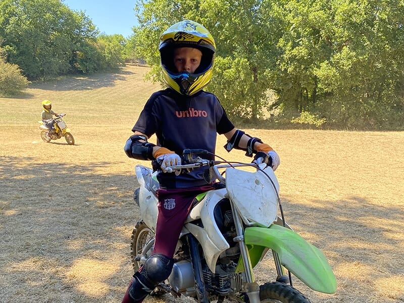 Jeune garçon portant un casque jaune et noir, assis sur une petite moto tout-terrain verte, sur un terrain sec et ensoleillé, avec un autre motocycliste à l'arrière-plan.