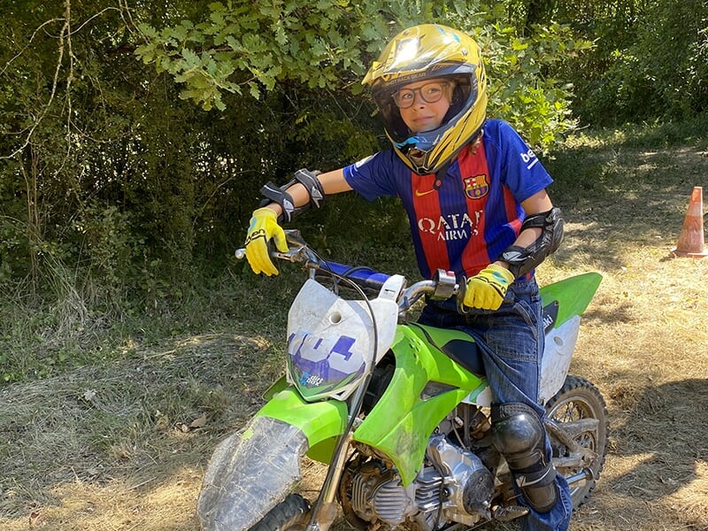 Jeune garçon avec des lunettes et un casque jaune, assis sur une moto verte tout-terrain, portant un maillot de football du FC Barcelone, à l'ombre d'arbres.