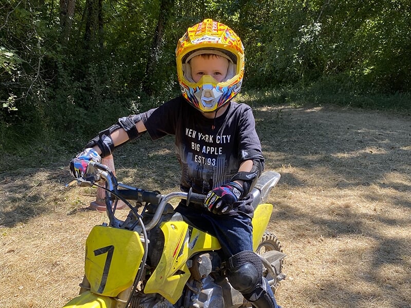Jeune enfant en casque jaune et orange, assis sur une moto tout-terrain jaune, sur un terrain sec à l'ombre des arbres.