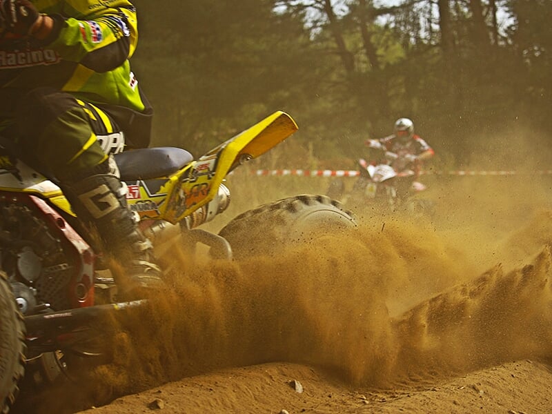 Vue rapprochée d'un quad jaune traversant un terrain sablonneux, créant un nuage de poussière avec un autre pilote en arrière-plan