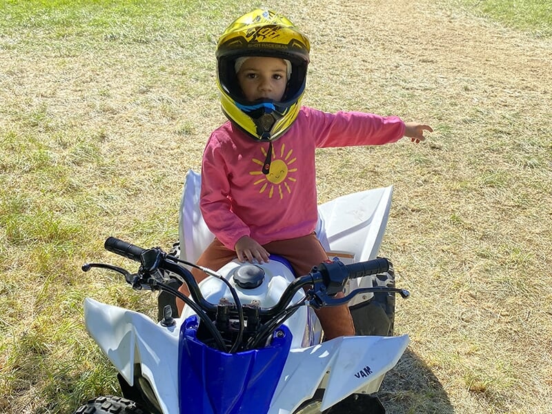 Un enfant portant un casque jaune assis sur un quad blanc, pointant du doigt sur le côté dans un environnement ensoleillé avec de l'herbe.