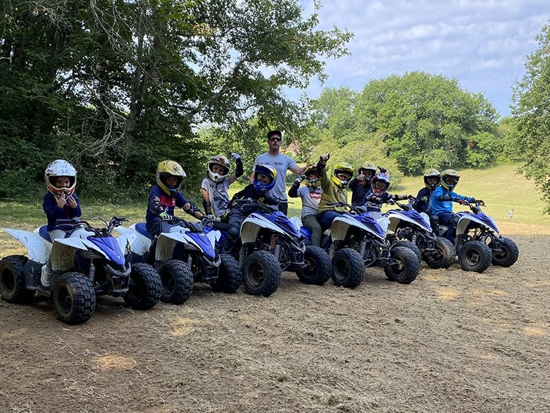 Un groupe d'enfants portant des casques et assis sur des quads alignés, accompagné d'un adulte, posant dans un cadre naturel avec des arbres et une prairie en arrière-plan.