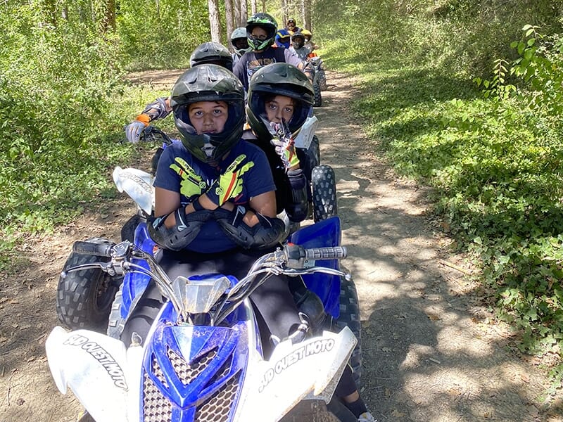 Un groupe d'adolescents ou de jeunes portant des casques noirs, assis sur des quads sur un chemin forestier, avec de la végétation dense autour.