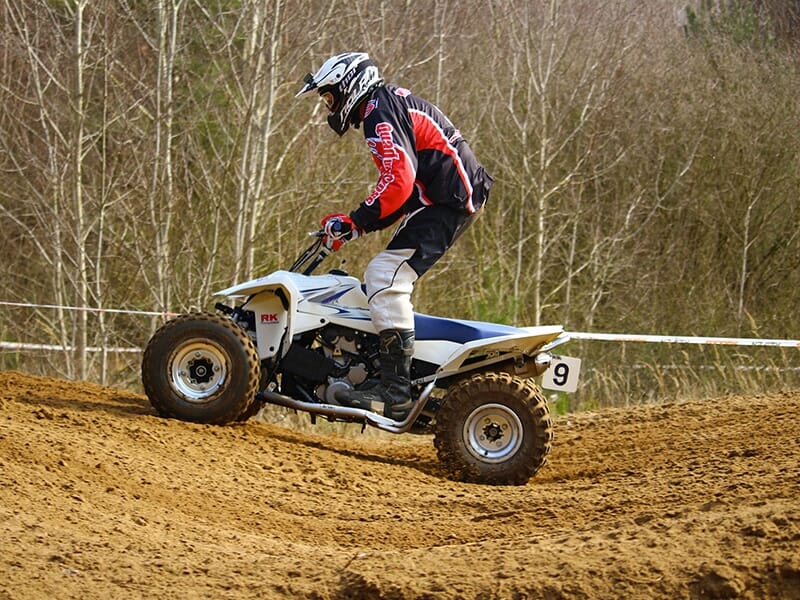 Un pilote portant un équipement de motocross, incluant un casque et une tenue rouge et noire, conduisant un quad blanc sur un terrain sablonneux avec des arbres nus en arrière-plan.