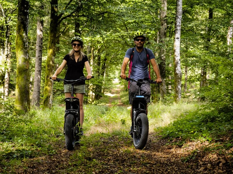 Un homme et une femme, tous deux en casque, roulant sur des trottinettes électriques tout-terrain dans un chemin boisé.