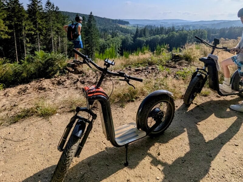 Une trottinette électrique tout-terrain garée sur un sentier avec vue sur une forêt vallonnée et un randonneur debout à l'horizon.
