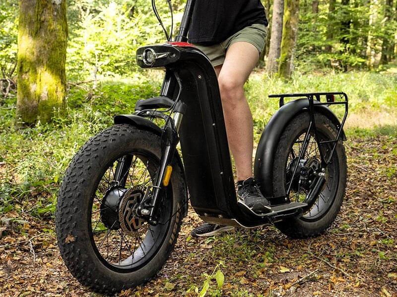 Un gros plan d'une trottinette électrique tout-terrain noire sur un sentier forestier, avec une personne dessus.