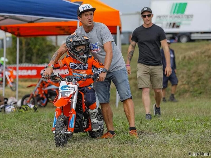 Enfant en tenue de motocross orange, aidé par un adulte, prêt à démarrer sa moto sur une pelouse.