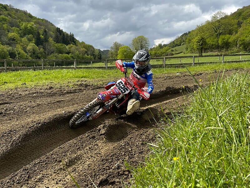 Motocycliste en tenue rouge et bleue dans un virage serré sur une piste boueuse, entouré de verdure.