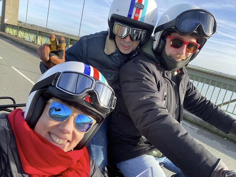 Trois personnes en casques et lunettes de protection, souriantes, dans une moto avec side-car noir, traversant un pont.