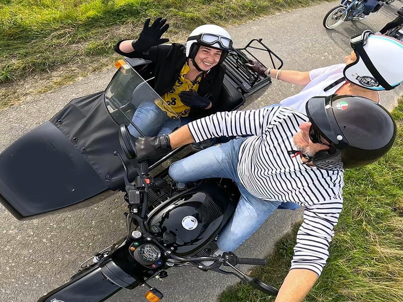 Prise de vue aérienne de deux personnes dans un side-car noir, l'un d'eux souriant et saluant, tandis que l'autre est à moto, roulant sur un chemin de campagne.