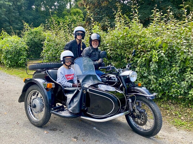 Trois personnes en casque, assises sur une moto avec side-car noir, stationnées près d'une haie de verdure, souriantes et prêtes pour une balade.