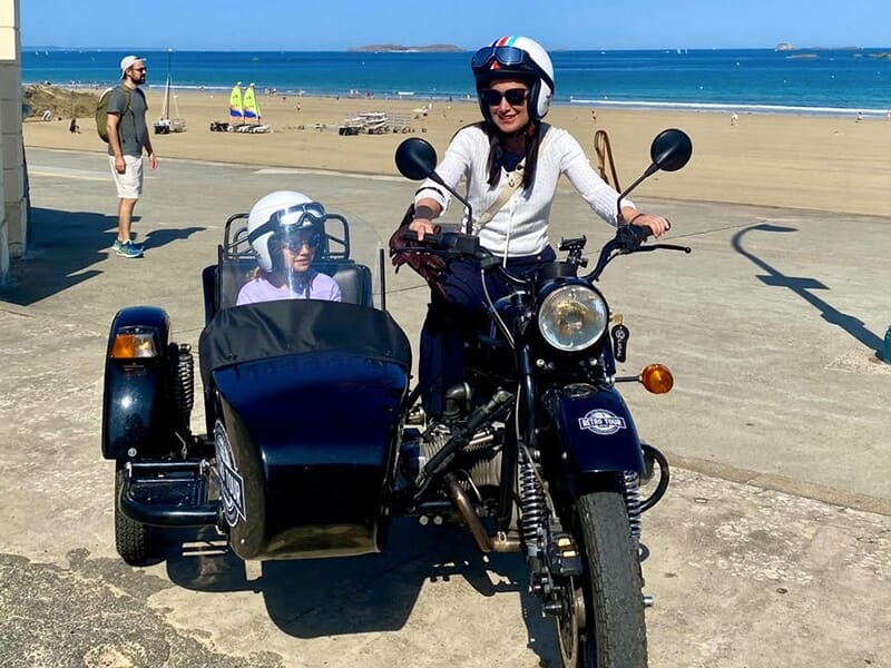 Une personne portant un casque de moto avec un passager dans un side-car, stationnés sur une promenade en bord de mer, avec une plage et des voiliers en arrière-plan.