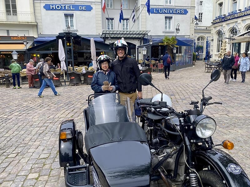 Deux personnes souriantes en casque, posant à côté d'une moto avec side-car noir garée sur une place pavée devant l'Hôtel de l'Univers, avec des terrasses de café et des passants en arrière-plan.