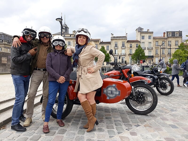 Groupe de quatre personnes portant des casques et lunettes rétro, posant devant des motos avec des side-cars, sur une place pavée avec des bâtiments historiques en arrière-plan.