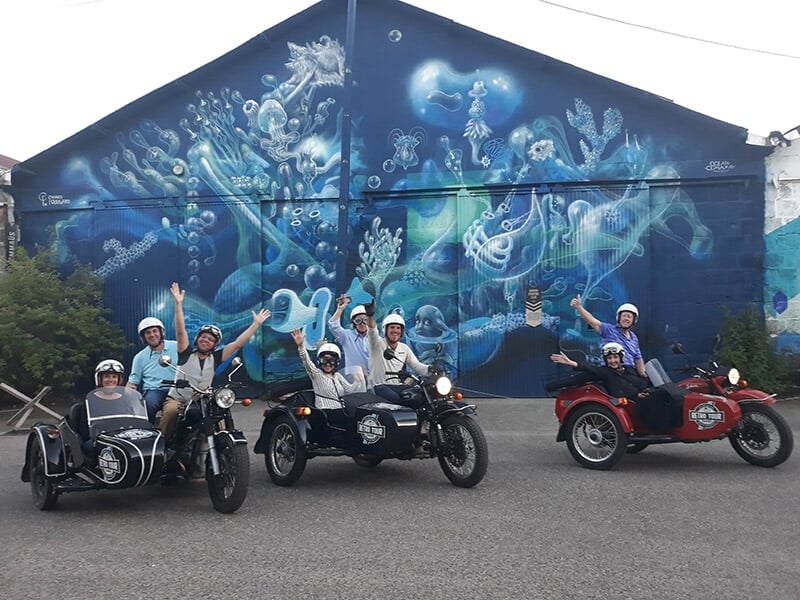 Groupe de huit personnes sur trois motos avec side-cars, bras levés, devant une fresque murale bleue représentant des formes marines et des créatures aquatiques.