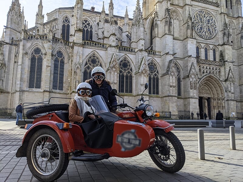 Deux personnes, casquées et assises dans un side-car rouge, posant devant une grande cathédrale gothique avec des détails architecturaux en arrière-plan.
