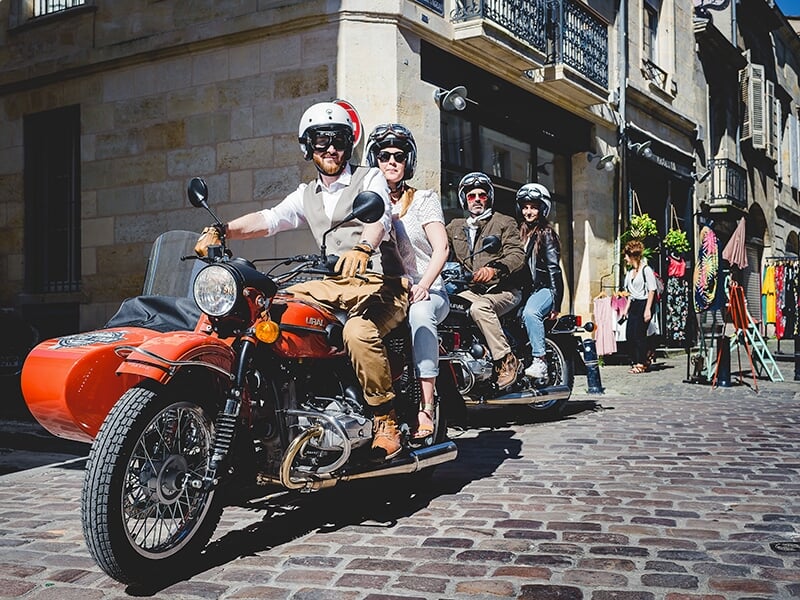 Deux motos avec side-cars alignées dans une rue pavée, chaque moto avec deux personnes casquées, posant dans un quartier historique avec des boutiques.