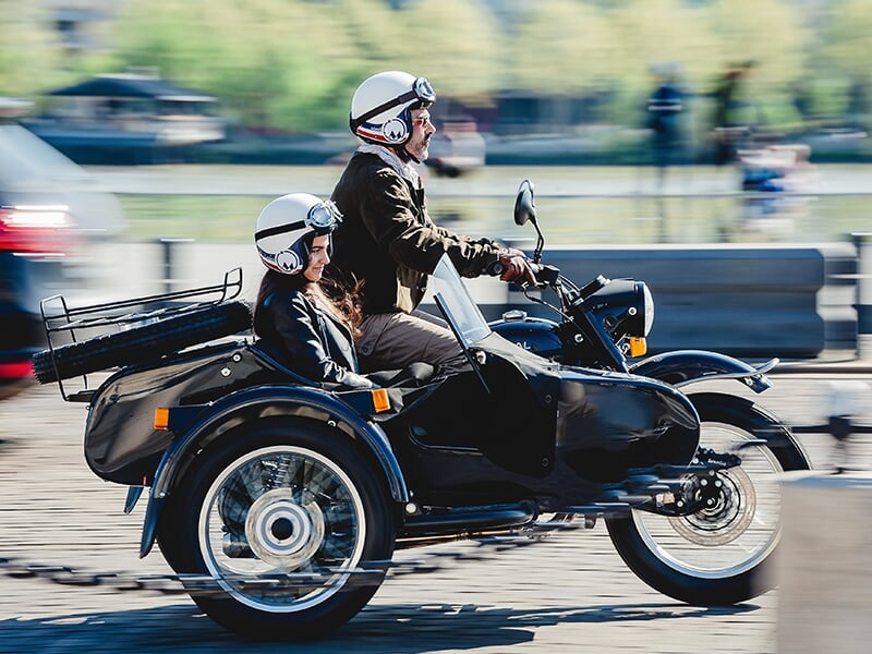 Homme conduisant une moto avec side-car noir, accompagné d'une femme, tous deux casqués, moto en mouvement sur une place publique.