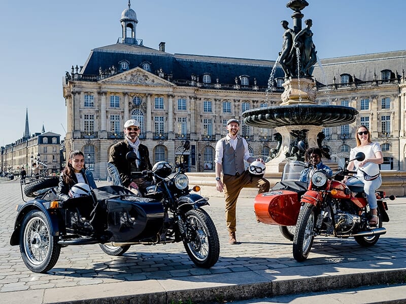 Deux motos avec side-cars, un homme debout entre elles, posant devant une grande fontaine historique dans une grande place publique.