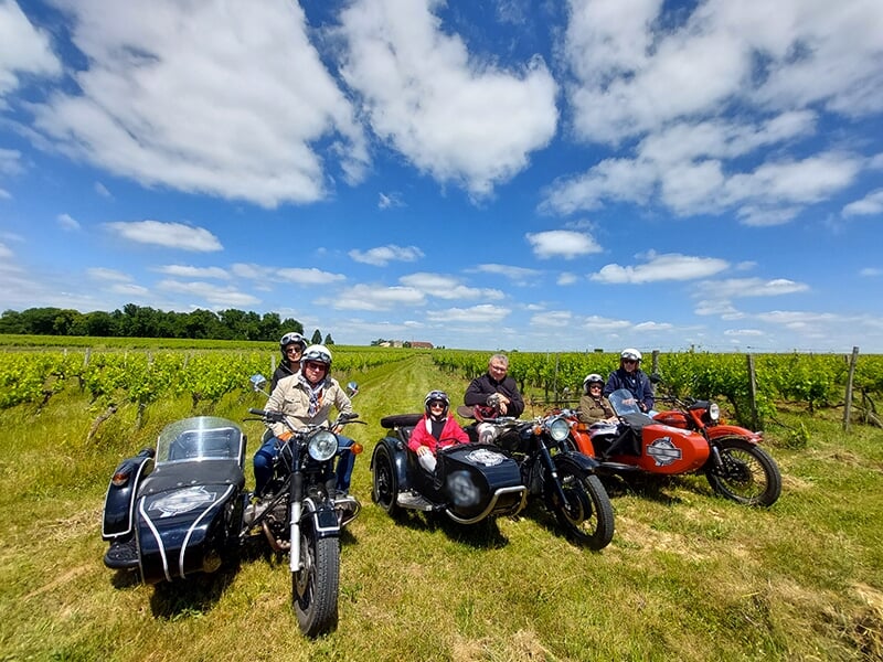 Groupe de personnes avec leurs motos et side-cars posant dans un vignoble sous un ciel ensoleillé avec quelques nuages.