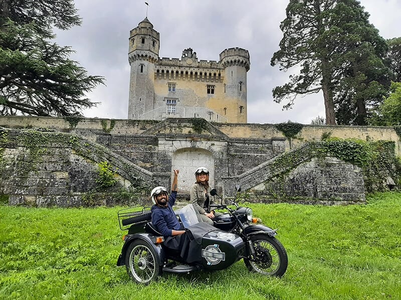 Deux personnes souriantes sur une moto avec side-car devant un château ancien entouré de verdure