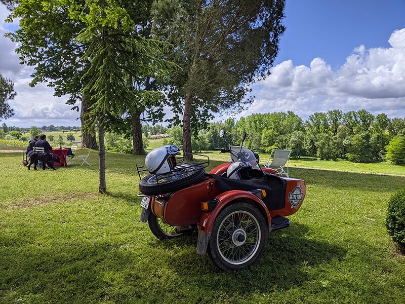 Une moto avec side-car rouge garée dans un champ sous un grand arbre, avec des tables de pique-nique en arrière-plan