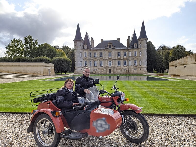 Deux personnes assises dans une moto avec side-car rouge devant un grand château avec des tours pointues.