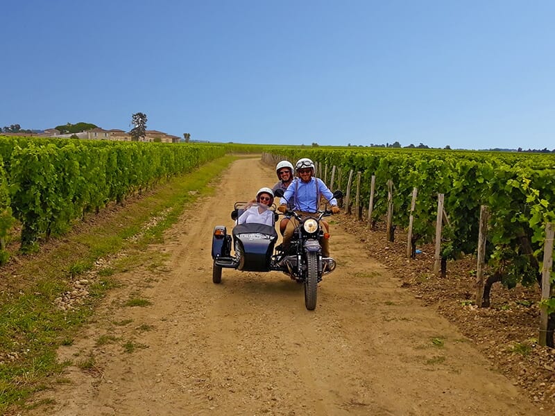 Trois personnes conduisant une moto avec un side-car dans un vignoble sous un ciel bleu