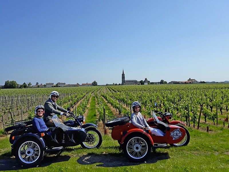 Deux motos avec side-cars dans un vignoble avec une église visible à l'horizon.