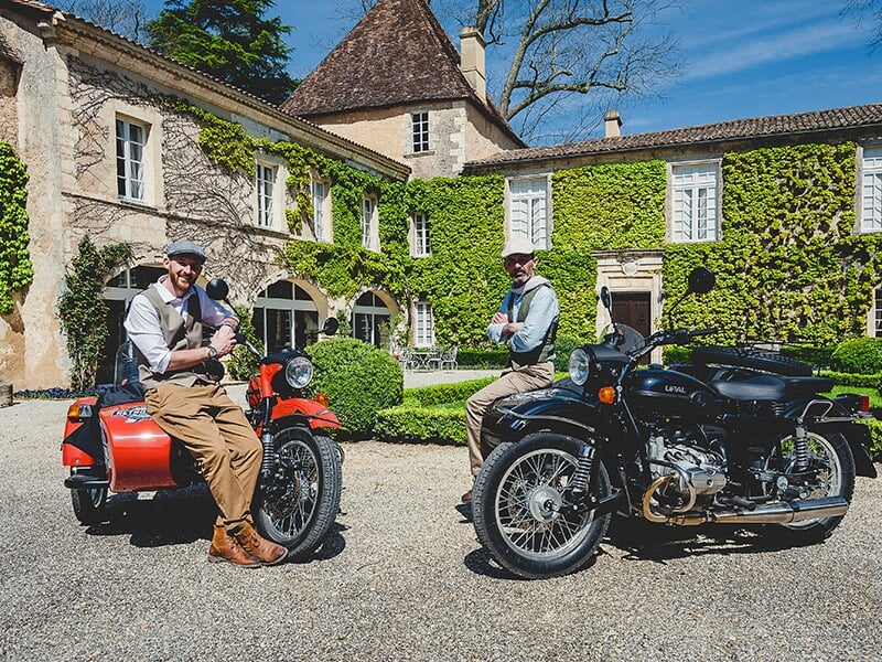 Deux conducteurs de motos avec side-cars posant devant une maison couverte de vigne.