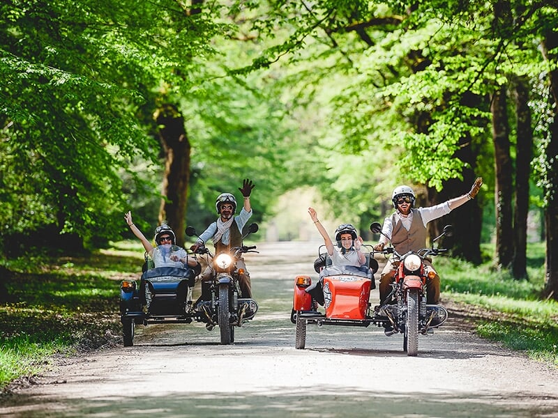 Deux motos avec side-cars, transportant des passagers enthousiastes, roulant sur un chemin forestier bordé d'arbres.