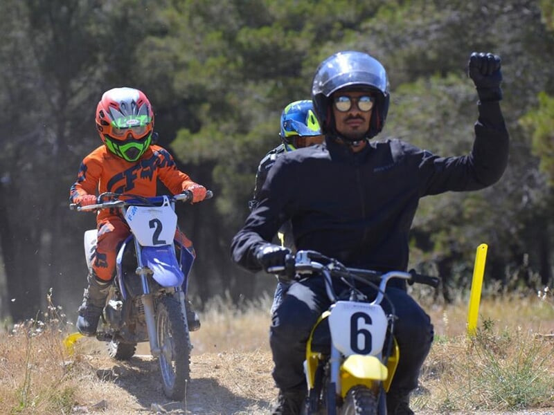 Pilote de moto portant un casque noir et levant le poing, suivi de jeunes motards sur un sentier en plein air.