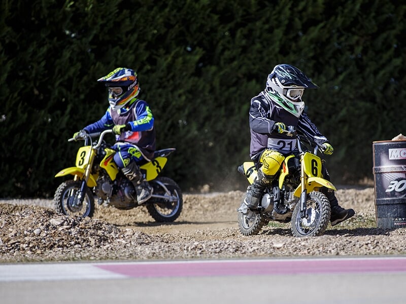 Deux jeunes pilotes de moto en tenue de course colorée, prenant un virage sur une piste en gravier.