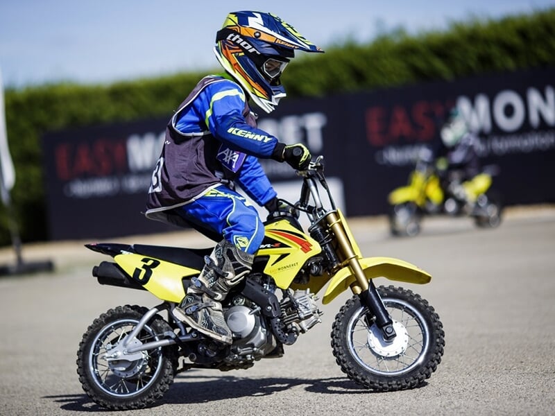 Enfant en tenue bleue et verte sur une petite moto jaune, roulant sur une piste en asphalte.