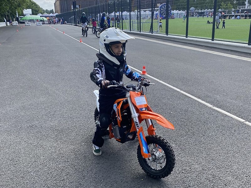 Enfant sur une moto orange, portant un casque blanc, roulant sur une route goudronnée délimitée par des cônes.