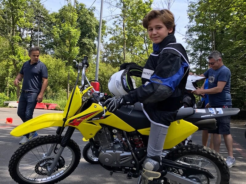 Adolescent souriant à côté de sa moto jaune, avec des adultes en arrière-plan, dans un environnement arboré.