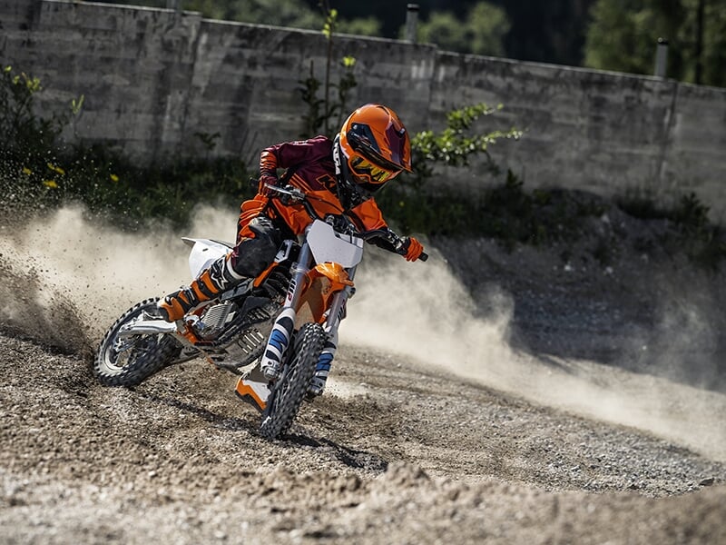Enfant en tenue orange vif et casque assorti, prenant un virage rapide sur un terrain poussiéreux.