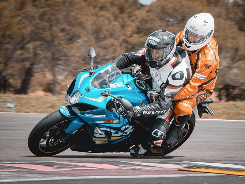 Motards en combinaison, l’un en orange et l’autre en noir, sur une moto bleue en pleine course sur circuit.